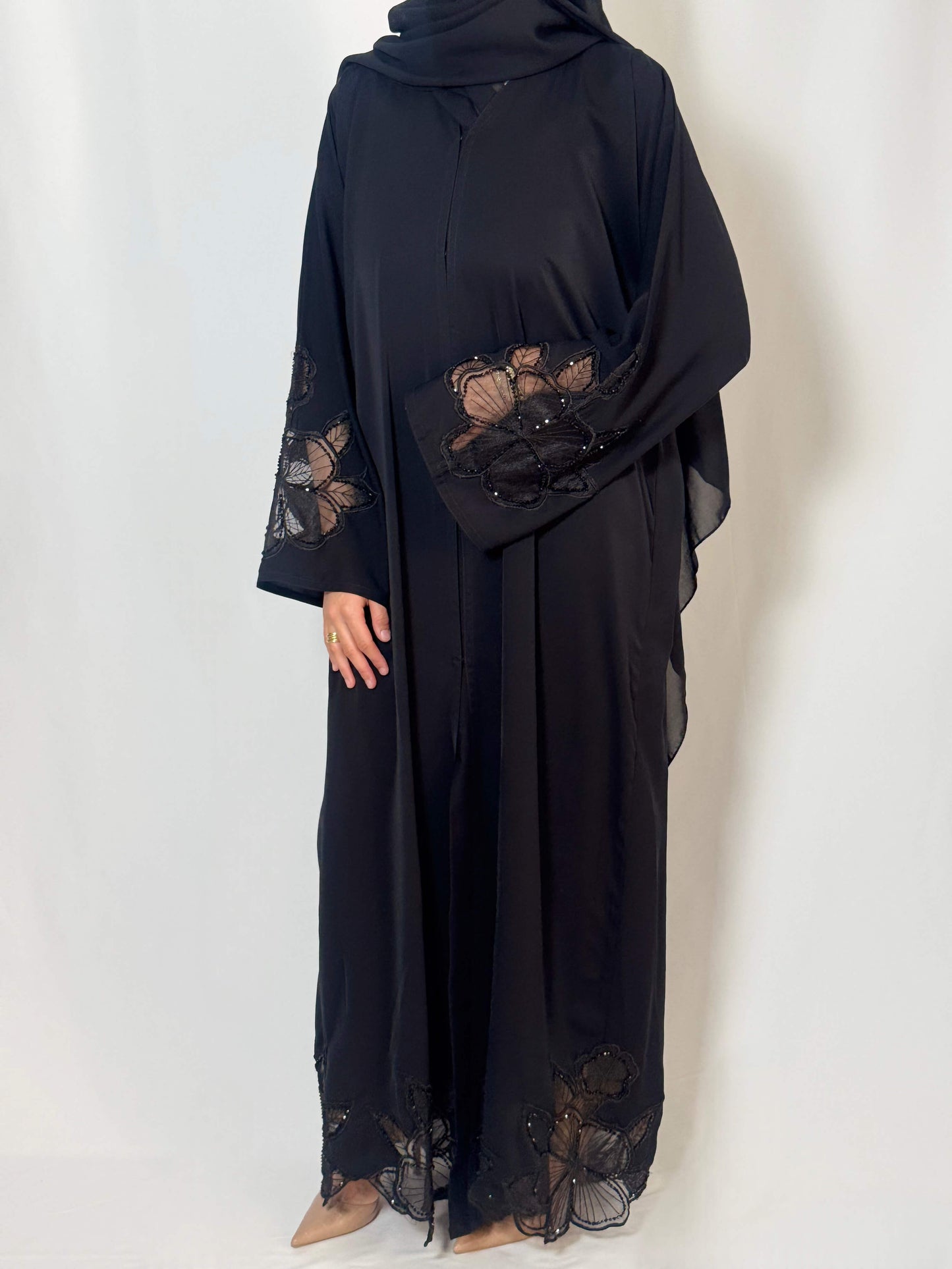 Abaya Camelia noir élégante portée par une femme voilée vue en entière silhouette fluide. légèreté des manches qui sont en dentelle transparente noire et ornées de perles noires qui brillent.