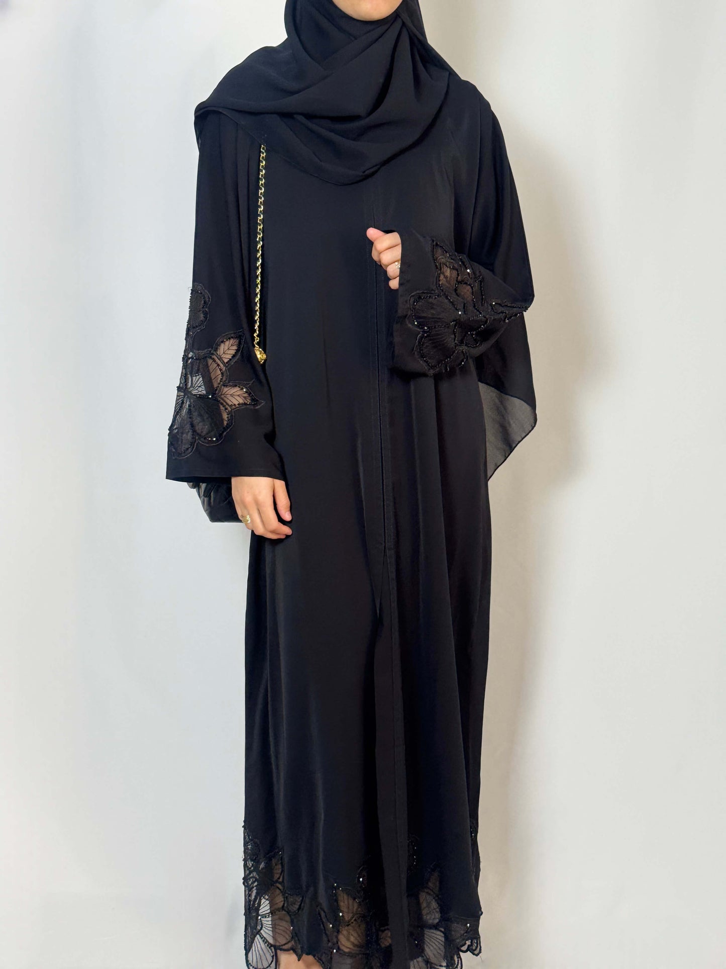 Abaya Camelia noir élégante chic et fluide en nidha. les manches sont en dentelle transparente noire et les fleurs sont ornées de perles noires qui brillent