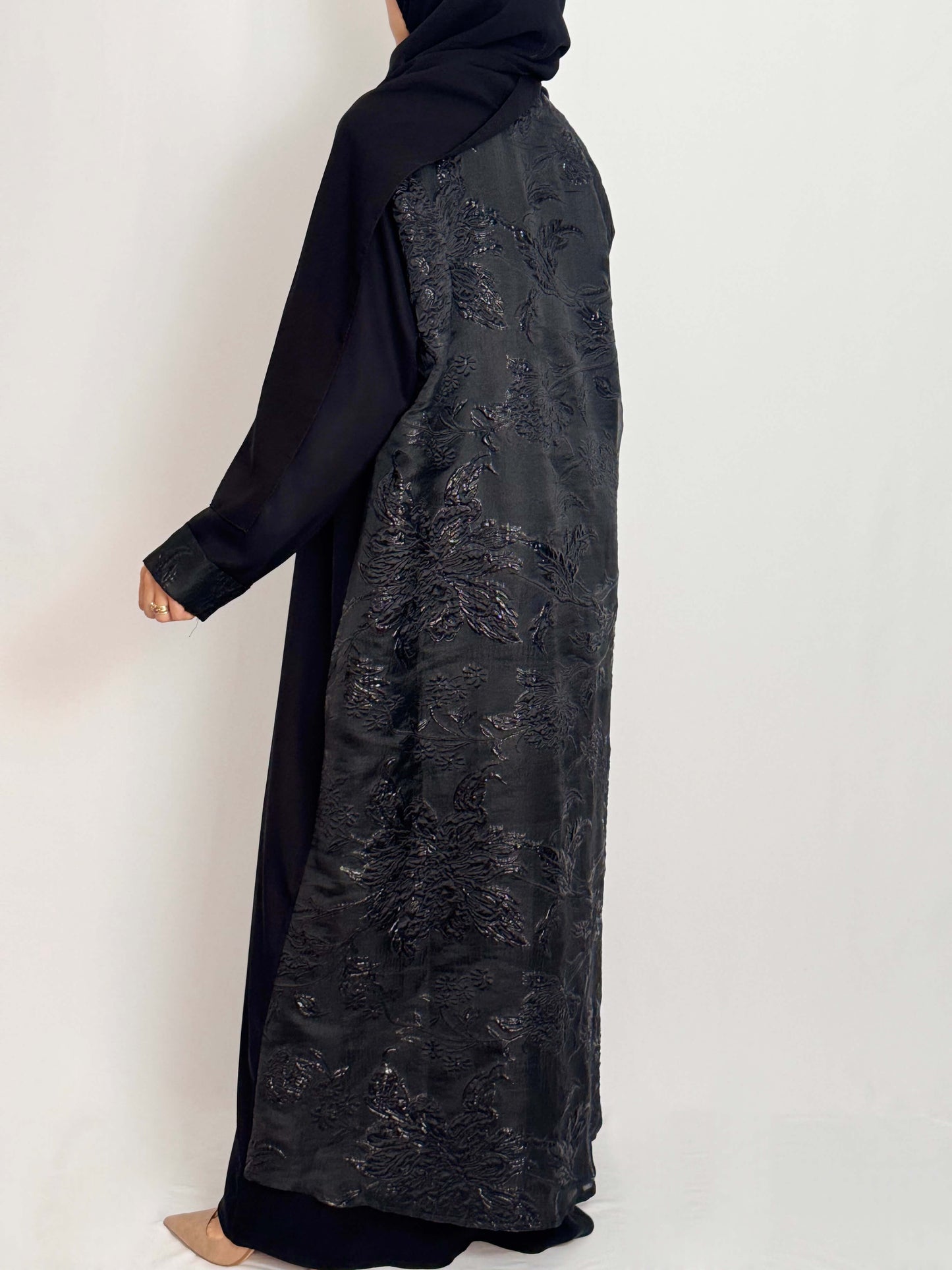 Abaya noire vue de dos portée par une femme voilé élégante silhouette aérienne en brocard irisé qui reflète la lumière