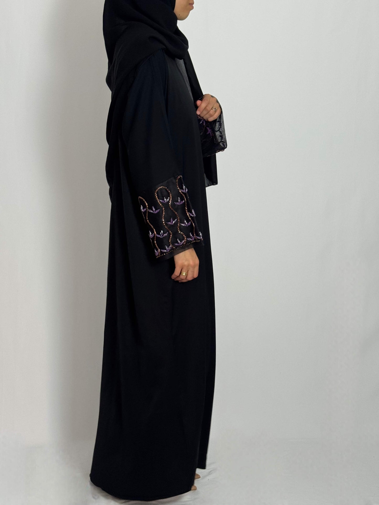 Abaya leila fluide et élégante tissus nidha vue de profil portée par une femme voilée sur fond blanc. abaya brodée aux manches avec des perleau couleur lila et rose