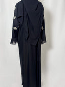 Abaya noire fluide en nidha vue de dos longue. les manches sont brodées de fleur argentées et un voile plissé apporte grace et élégance aux bout des manches 