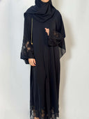 Abaya Camelia noir élégante chic et fluide en nidha. les manches sont en dentelle transparente noire et les fleurs sont ornées de perles noires qui brillent 