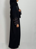 Abaya leila fluide et élégante tissus nidha vue de  profil portée par une femme voilée sur fond blanc. abaya brodée aux  manches avec des perleau couleur lila et rose 